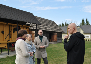Četri pieauguši cilvēki sarunājas muzeja sētā pie senlaicīga labības kulšanas kombaina