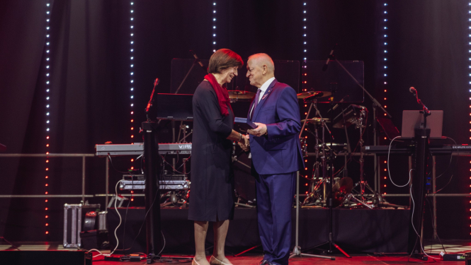 Sieviete un vīrietis viens otram paspiež roku. Vīrietis pasniedz Dobeles novada pašvaldības apbalvojumu