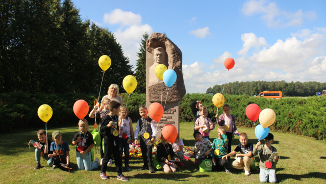 1. klases skolēni ar krāsainiem baloniem sasēduši pie K.Ulmaņa pieminekļa muzejā "Pikšas"