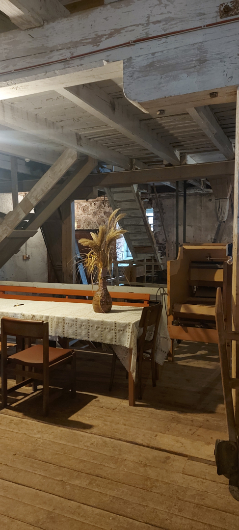 Telpa dzirnavās, kurās ir divas koka kāpnes uz nākamo stāvu un liels galds ar krēsliem un puķupodu uz tās