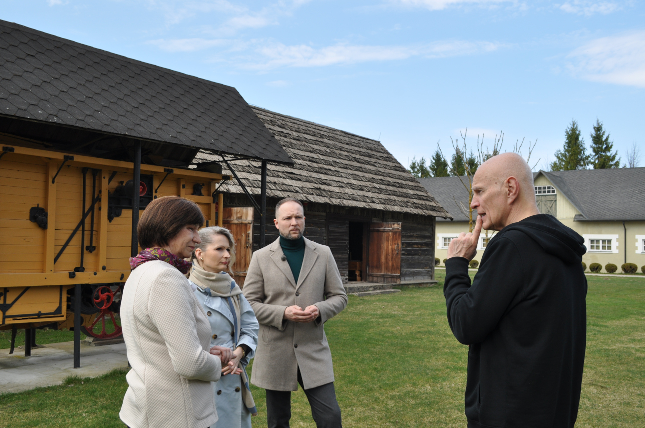 Četri pieauguši cilvēki sarunājas muzeja sētā pie senlaicīga labības kulšanas kombaina